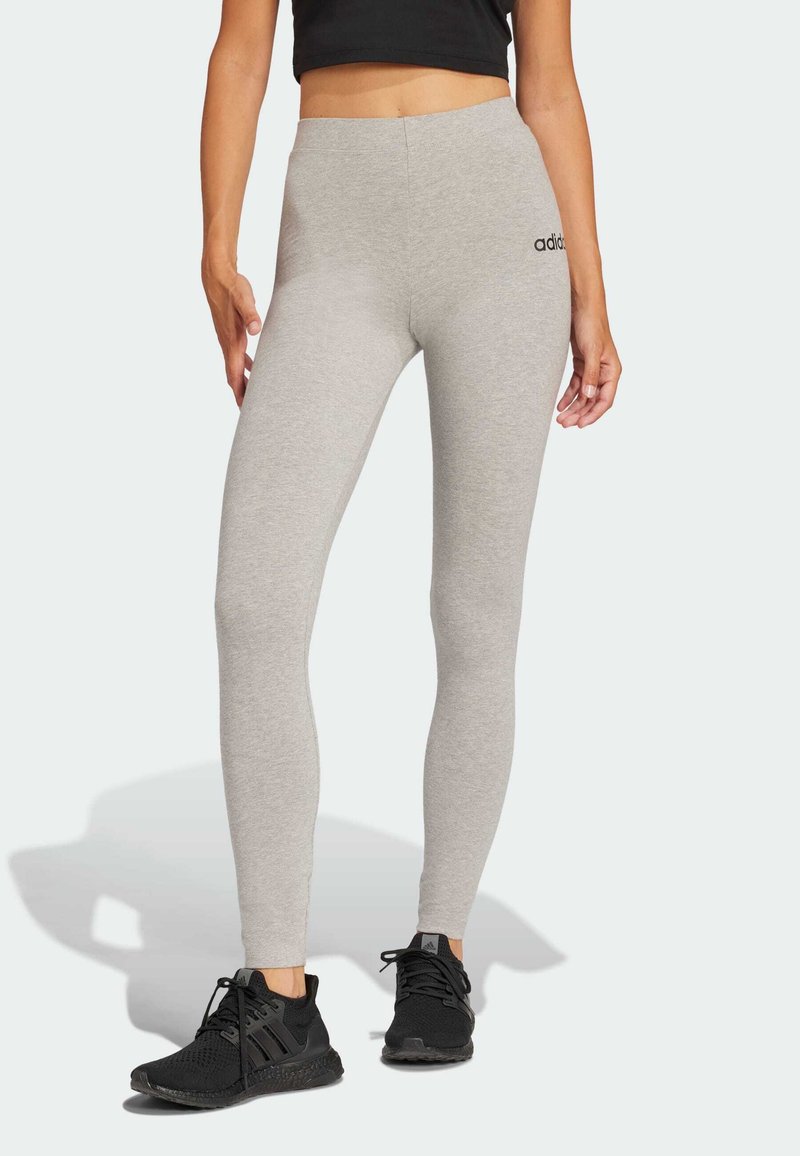 Szürke leggings, puha anyagból készült, magas derékkal és fekete Adidas logóval a csípőjén. Fekete sportcipővel párosítva.