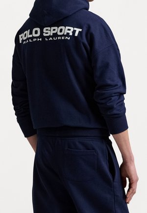 Personne portant un sweat à capuche bleu marine et un pantalon assorti avec l'inscription "Polo Sport Ralph Lauren" en blanc au dos du sweat.