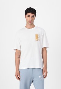 Hvid bomulds t-shirt med korte ærmer, prydet med et lysbrunt logo design på venstre brystområde. Behagelig, afslappet pasform.