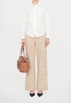 Une personne porte une chemise blanche boutonnée, un pantalon cargo beige à jambes larges, des talons noirs pointus, et tient un grand sac à main marron par les anses.