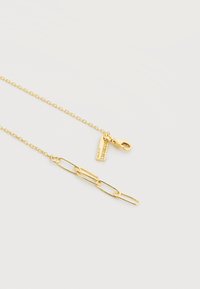 Χρυσό κολιέ αλυσίδα με κούμπωμα αστακού και μια μικρή ορθογώνια ταμπέλα χαραγμένη με τη φράση "ΚΑΤΙ ΑΛΛΟ".