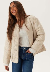 Beige gewatteerde jas met een boxy ontwerp, voorzien van knoopsluitingen en textielstof. Gecombineerd met een witte top en hoge taille denim jeans.
