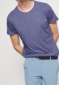 Lila Heidekraut T-Shirt mit kurzen Ärmeln, Rundhalsausschnitt und kleinem Logodetail auf der Brust, kombiniert mit hellblauen Hosen und einem schwarzen Gürtel.