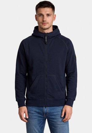 Młody mężczyzna ubrany w granatową bluzę z kapturem na zamek z przednimi kieszeniami oraz jasnoniebieskie jeansy, stojący na tle jednolitego białego tła.