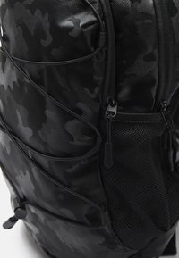 Zaino camouflage nero con tessuto testurizzato, design curvo, multiple cerniere, dettagli in rete e tracolle regolabili.