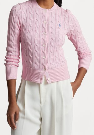 Femme portant un cardigan boutonné à manches longues en maille torsadée rose clair et un pantalon blanc taille haute à jambes larges.