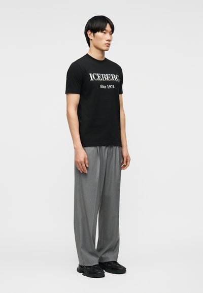 Νεαρός άνδρας όρθιος, φορώντας μαύρο T-shirt με την επιγραφή "ICEBERG since 1974", γκρι φαρδιά παντελόνια και μαύρα αθλητικά, με φόντο ένα απλό χρώμα.