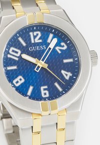 Orologio in acciaio inossidabile argento e oro con quadrante blu texturizzato, numeri bianchi e un accento dorato sulla lancetta del secondo. Movimento al quarzo.