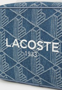 Borsa in denim con un motivo geometrico blu e grigio, con la scritta bianca "LACOSTE" e "1933" ben in evidenza.