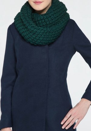 Femme portant une écharpe infinie en tricot épais de couleur teal foncé et un manteau bleu marine, la main posée sur la hanche.