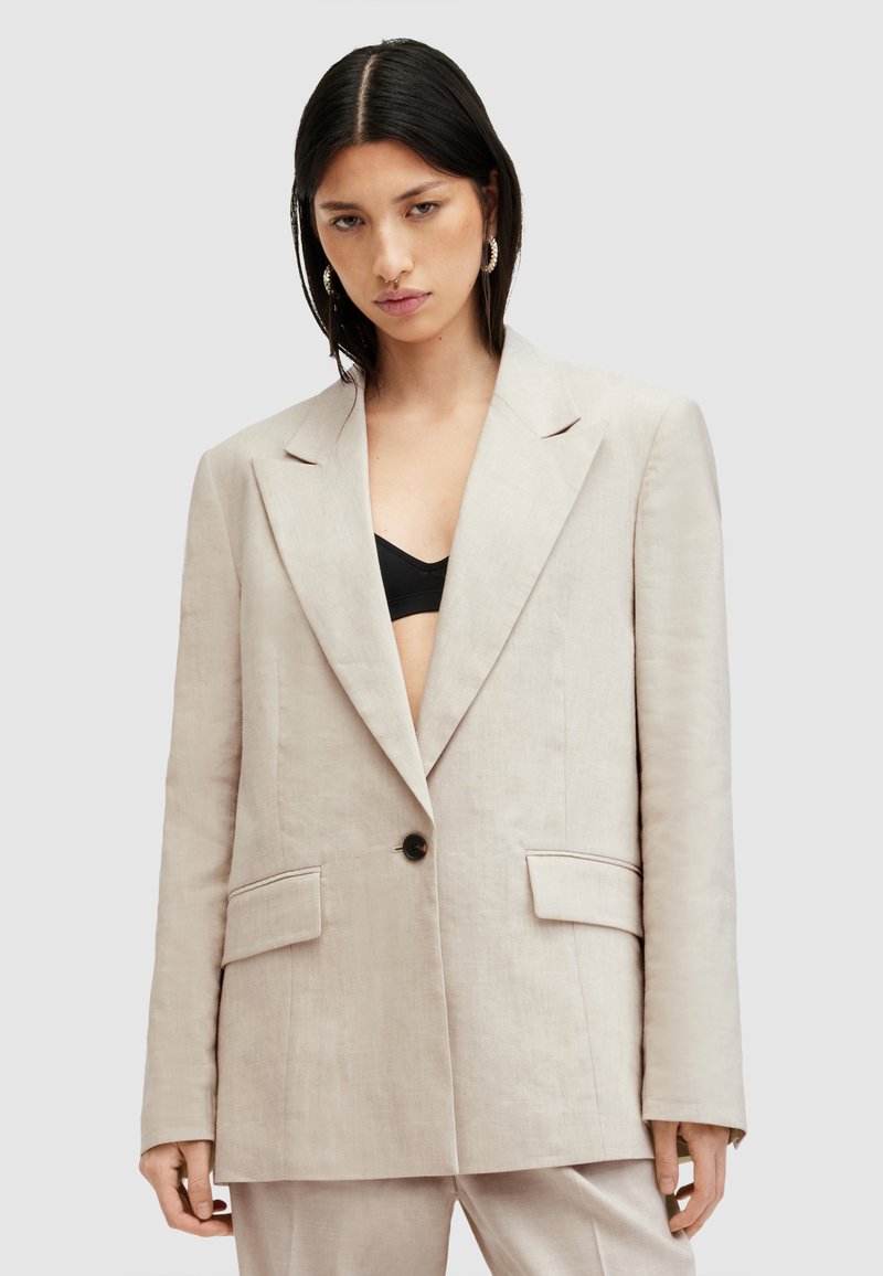Blazer in lino beige con un design strutturato, ampi revers, chiusura con un solo bottone nero e due tasche frontali. Stile minimalista.