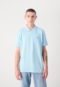 Lyseblå polo T-shirt med korte ærmer, tre-knaps placket og et lille logo på brystet. Et par lyseblå jeans bæres.