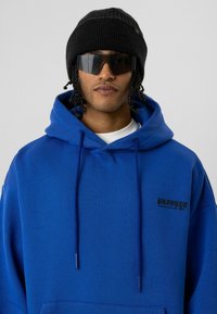 Blauwe hoodie met trekkoorden, voorzak en zwarte geborduurde tekst. Draag met een zwarte gebreide beanie en donkere zonnebril. Gladde textuur.