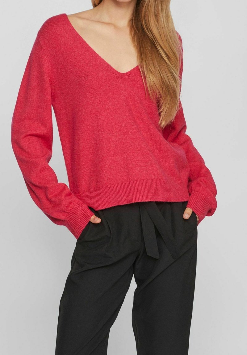 Femme portant un pull rouge ample à col en V avec manches longues et un pantalon noir taille haute avec une ceinture nouée, mains dans les poches.