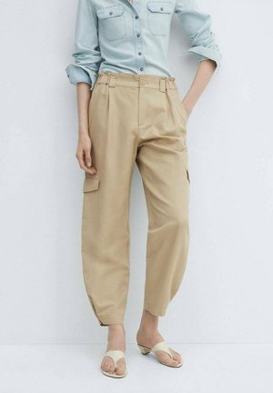 Vrouw in een lichtblauw hemd met knopen, ingestopt in beige cargobroek en witte sandalen met hakken, staand tegen een effen achtergrond.