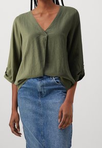 Olivgrüne, leichte Wickelbluse mit tiefem V-Ausschnitt und Dreiviertelärmeln, kombiniert mit einem hellblauen Denimrock.