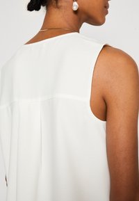 Haut blanc sans manches avec une couture d'empiècement dos et un pli, fabriqué dans un tissu lisse et léger, avec une coupe décontractée.