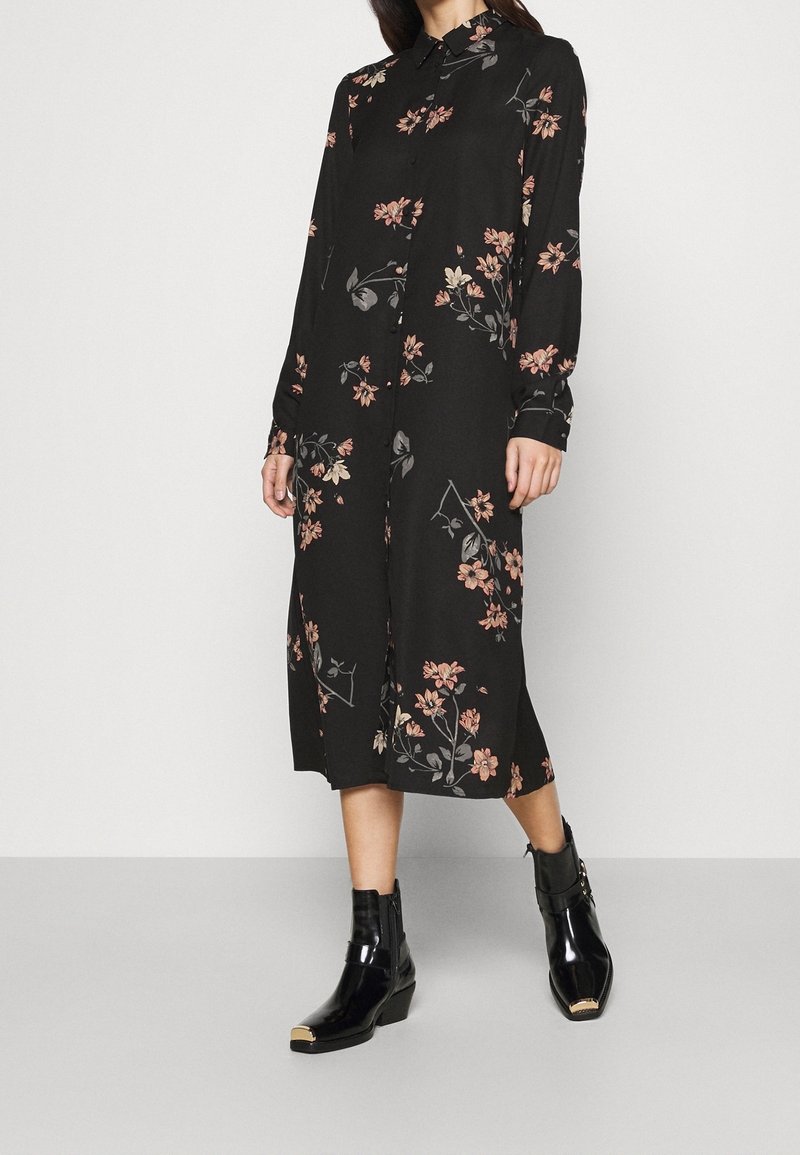 Femme portant une robe noire mi-longue ornée d'un imprimé floral rose et à manches longues, assortie à des bottines noires avec embouts dorés.