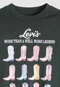 Μαύρο μπλουζάκι με πολύχρωμα γραφικά καουμπόικων μπότες σε παστέλ αποχρώσεις. Το λογότυπο και το σύνθημα της "Levi's" τυπωμένα πάνω από τις μπότες σε λευκή τυπογραφία.