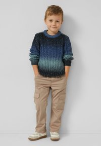 Gruby sweter z dzianiny o gradiencie od ciemnoniebieskiego do jasnoturkusowego, zestawiony z beżowymi spodniami typu cargo i białymi trampkami.