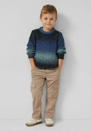 Gruby sweter z dzianiny o gradiencie od ciemnoniebieskiego do jasnoturkusowego, zestawiony z beżowymi spodniami typu cargo i białymi trampkami.