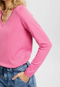 Różowy sweter z dzianiny z dekoltem w serek i długimi rękawami. Materiał wydaje się miękki i gładki. Zestawiony z niebieskimi dżinsami, prezentując casualowy styl.