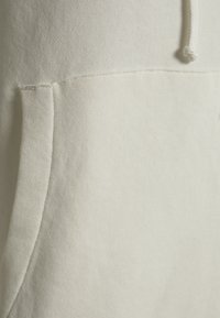 Beige fleece-stoftekstur med synlige syninger, med en frontkangaroo-lomme og snorehardware på hætten.