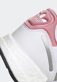 Vitaid sportskor i vitt med rosa accenter, slät syntetisk material, texturerad sula och silverdetaljer nära hälen. Har en logotyp på baksidan.