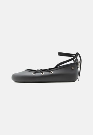 Black matte ballet flat with lace-up front, ankle wrap tie, and small metallic logo charm at heel.