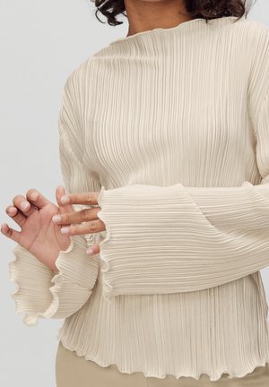 Femme portant un haut plissé à manches longues couleur crème avec des poignets et un ourlet volantés, ajustant les manches avec ses mains visibles.