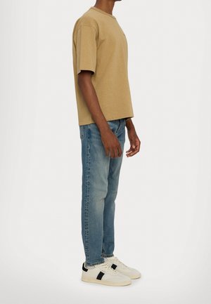 Person trägt ein beige kurzärmeliges Hemd, blaue Jeans und weiße Sneaker mit schwarzen Streifen und steht seitlich vor einem einfarbigen Hintergrund.