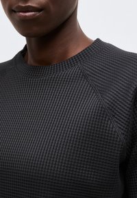 Κοντινό πλάνο ενός ατόμου που φοράει σκούρο γκρι, υφασμένο πουλόβερ με υφή βάφλας και στρογγυλή λαιμόκοψη, εμφανίζοντας τον λαιμό και το πάνω μέρος του στήθους.