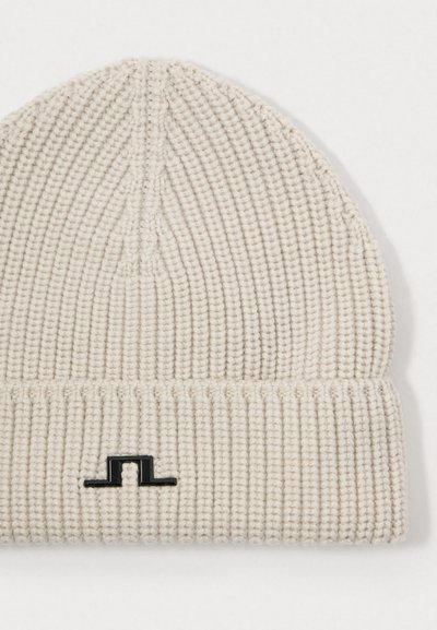 Knitted beanie in light beige, featuring ribbed texture. Black logo accent on the cuff. Close-up view showing material details.