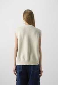 Ärmelloses Stricktop in Creme mit gerippter Textur, ausgestattet mit einem runden Ausschnitt und einem taillierten Saum, getragen mit dunklen Jeanshosen.