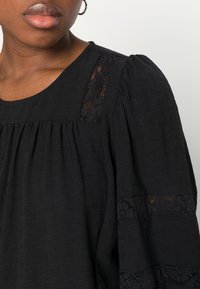 Blusa preta com mangas bufantes, apresentando detalhes em renda nos ombros e um frente plissada. Tecido texturizado, decote redondo.