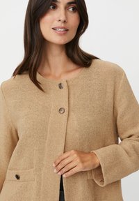 Beige strukturierte Jacke mit rundem Ausschnitt, zwei Vordertaschen und einem Verschluss mit dekorativem silberfarbenem Knopf.