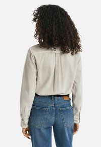 Ljusbeige långärmad skjorta med plissédetaljer på baksidan, tillsammans med medelblå denimjeans med dubbla bakfickor.