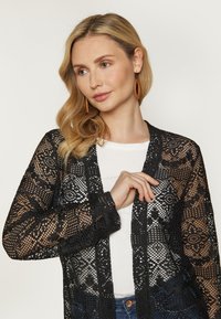 Cardigan in pizzo nero con intricate fantasie floreali, design aperto, maniche lunghe e polsini a coste, indossato sopra una maglia bianca.