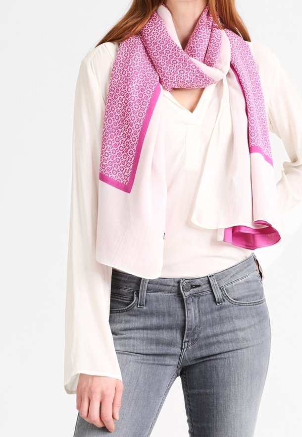 Femme portant une écharpe rose clair avec un motif géométrique rose sur un chemisier blanc et un jean gris sur fond blanc.