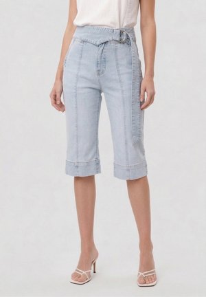 Kvinde iført lyseblå knælange denimshorts med bred talje og bælte-detalje, kombineret med hvide, rem-højhælede sandaler.