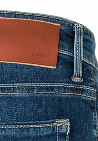 Dunkelblaue Denim-Jeans mit kontrastierenden orangen Nähten. Besitzt ein braunes Lederetikett mit der Aufschrift "MAC JEANS" am Bund.