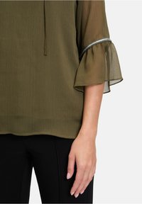 Blusa a coste color verde oliva con maniche a campana trasparenti. Presenta un bordo argentato lungo il lato delle maniche, una texture liscia e una vestibilità casual.