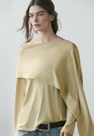 Giovane donna con lunghi capelli, indossa un top asimmetrico giallo chiaro, cintura nera e jeans blu su uno sfondo chiaro e uniforme.