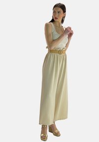 Beige wijde broek met elastische taille en ruches, gecombineerd met een aansluitend crèmekleurig hemdje en gouden platformsandalen.