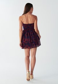 Mouwloze, gelaagde jurk in donkerrood en marineblauw met bloemenpatroon, een smock-borstvoeding, doorzichtige stof, gecombineerd met gouden sandaaltjes.