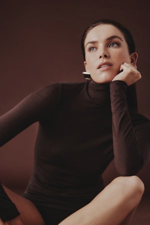 Strak aansluitende bruine coltrui met lange mouwen, gladde textuur, gecombineerd met zilveren oorbellen. Achtergrond in warme bruintinten.