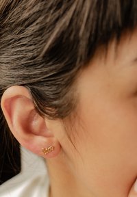 Pendiente de aro dorado con forma de la palabra "amor", presenta una superficie suave y un diseño delicado, llevado en un lóbulo con cabello oscuro.