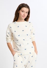 Sudadera color crema hecha de una tela suave con bordado de lazo azul en toda la prenda. Mangas largas con un escote redondeado y un corte relajado.