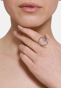 Anello in metallo tono argento con un design fluido e intrecciato. Texture liscia e forma minimalista, posizionato su un dito vicino al mento.