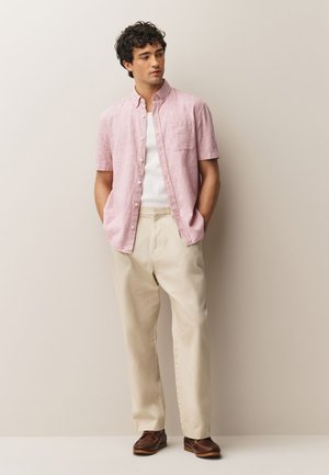 Mladić stoji s rukama u džepovima noseći svijetloružičastu košulju kratkih rukava na kopčanje, bijelu potkošulju, bež hlače i smeđe cipele.